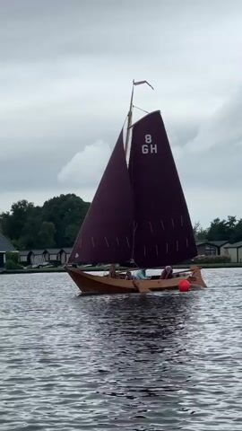 A sailboat with two sails, one maroon and one dark, is sailing on a body of water. The sails have the number 8 and the letters GH printed on them. A few people are visible on the boat. The boat is moving from left to right across the frame. A red buoy is visible in the water to the right of the boat. In the background, there are trees and buildings that appear to be houses or cabins. The sky is cloudy and overcast. The water is choppy, with ripples and waves. The timestamp indicates the video is approximately 4 seconds long.