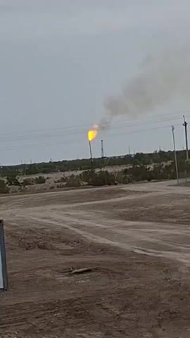 The video shows a distant flare stack burning gas against an overcast sky. In the foreground, a dirt road with tire tracks leads towards the flare stack. The area around the road appears to be arid with sparse vegetation. Power lines and utility poles are visible in the mid-ground. The overall scene suggests an industrial or oil and gas extraction site in a dry, possibly remote location. The camera angle is low, looking up towards the flare stack.