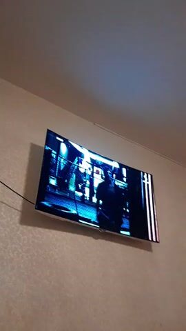 The video shows a close-up of a television screen displaying a scene from a movie or TV show. The screen is mounted on a textured wall, and a black cable is visible running from the TV. The content on the screen features a man in a light-colored shirt, with a backdrop that appears to be an urban environment with fencing and lights. The lighting suggests it might be evening or nighttime. The man's expression is not clearly discernible due to the image quality and the angle. There is some text or a logo in the top right corner of the screen, but it is too blurry to read. The overall scene implies someone is watching content on their TV.