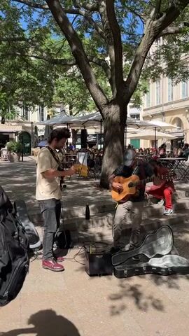 Here is a description of the video:

The video shows two street performers playing guitars in a European town square. One is playing an acoustic guitar, while the other has an electric guitar. They are standing in the shade of a large tree, and there are several open guitar cases on the ground in front of them.  Behind them are outdoor cafes with white umbrellas, tables and chairs, and people sitting and enjoying the sunny day.  A man sits on the steps behind them with his legs crossed. A small amplifier sits on the ground.