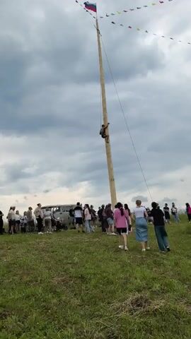 A crowd of people watches a man climb a tall wooden pole. The man is midway up the pole, gripping it with his hands and knees. He is wearing a dark t-shirt and shorts. A rope is attached to the top of the pole and extends down towards the man, possibly for assistance or as part of a competition. The pole is decorated with small flags of various colors at the very top, and a Russian flag is also visible at the summit. The people below are standing on a grassy hill, some are casually dressed, and some are observing the climber intently. In the background, a large vehicle resembling a van is parked, and the sky is overcast with dramatic clouds. The overall scene appears to be an outdoor event or festival.