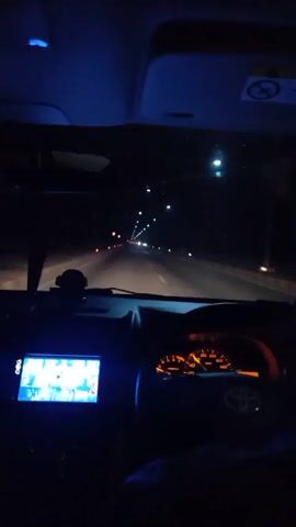 The video shows the interior of a car at night, from the perspective of the driver. The dashboard is visible, with a screen displaying what appears to be a navigation system or media player, and the speedometer gauge illuminated with an orange glow. The windshield shows a dark road ahead, lined with streetlights and other vehicle lights receding into the distance. The ambient light is mostly from the car's interior and the external lighting of the road.