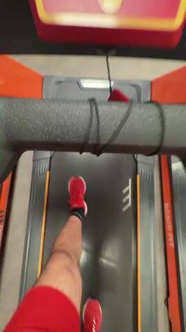 A first-person perspective video shows a person running on a treadmill. The person is wearing red athletic shorts and red running shoes. The treadmill is a MATRIX brand and has an orange and black color scheme. The display shows the time as 10:36 and then 10:37. The person's legs are in motion, indicating they are running. The environment appears to be a gym, with other treadmills visible in the background. A safety clip is attached to the person's shorts and to the treadmill. The video is short and focuses on the act of running.