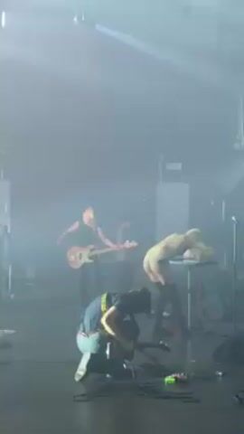 The video shows a performance on a stage. The stage is filled with fog and there are flashing lights. There are at least three people on the stage. One is wearing a yellow outfit and is bending over a small table. Another is wearing dark clothing and is kneeling on the stage, possibly adjusting equipment. A third person is standing with a guitar. The lighting changes frequently, obscuring the view and making it difficult to see details. The atmosphere appears to be energetic and intense.