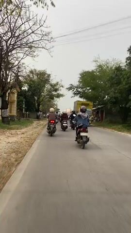 The video shows a group of people riding motorcycles on a paved road. The road appears to be in a rural or semi-urban area with trees and buildings on either side. The sky is overcast. The motorcyclists are wearing helmets and casual clothing. Some of the motorcycles are carrying passengers. A truck is also visible on the road. The video is shot from the perspective of someone riding a motorcycle within the group, looking forward.