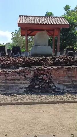 The video pans across an outdoor archaeological site on a sunny day. In the foreground is a dirt path, followed by a low wall constructed of rough stones. Behind this, a larger wall made of red bricks is in ruins, with a significant section of the wall crumbling inwards, exposing a pile of fallen bricks. Atop this ruined brick wall are several dark, rectangular stone structures. In the background, under a wooden shelter with a tiled roof, sits a large, light-colored, roughly shaped stone or monument. Trees and foliage are visible behind the shelter, and the sky is clear and blue. The camera movement is slow and steady, offering a detailed view of the ancient structures. There are no people visible in the video.