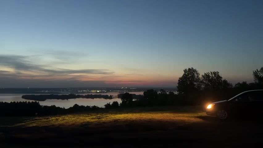 The video shows a landscape at twilight. The sky is a gradient of blues and pinks, indicating a recent or impending sunset. Below the horizon is a body of water with visible islands and what appears to be a city's lights in the distance. The foreground features a grassy hill, with a car parked on the right side, its headlights illuminating the immediate ground. Trees are silhouetted against the water and sky. The scene is still and quiet.