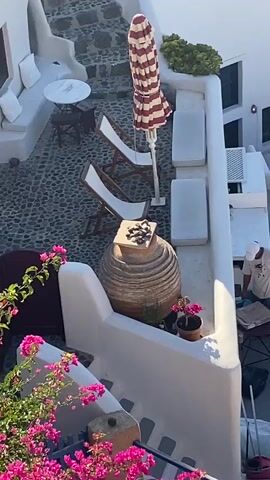 The video pans over a white-washed village built on a cliff overlooking a blue ocean. In the foreground, a person in a light-colored shirt and white cap is working, bent over a table. Several white chairs and umbrellas are visible on patios. The architecture is distinct, with curved white walls and staircases. In the background, the vast expanse of the ocean stretches to the horizon, dotted with a few sailboats. Mountains are visible in the far distance. The overall scene is sunny and picturesque, suggesting a Mediterranean or Greek island setting.