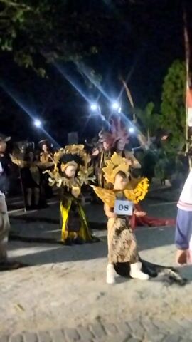 This video captures a nighttime outdoor event, likely a parade or performance, featuring individuals in elaborate costumes. The focus is on several performers, each adorned with distinctive, large headpieces resembling sunflowers or sunbursts, embellished with gold and brown elements. One performer, with the number 08 prominently displayed on a white card, stands out. The lighting is dim, with string lights illuminating the scene and creating lens flare effects. The background suggests an urban or public space with some greenery and a paved surface. Several other figures are visible in the background, some also in costumes, and a crowd appears to be observing the event. The overall atmosphere is festive and visually striking due to the vibrant costumes against the dark backdrop.