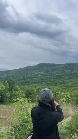 A person wearing a grey baseball cap and a black jacket is standing outside, facing away from the camera and toward a green, forest-covered mountain. The person holds up a mobile phone to take a picture of the mountain. The sky is filled with grey clouds. The foreground features shrubs and a dirt path. The mountain fills the horizon, covered densely with trees. The overall mood is one of contemplative observation in a natural setting.