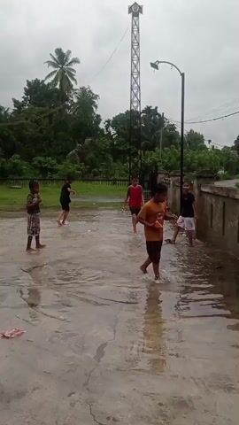 The video shows several children playing in a flooded courtyard on a cloudy day. The ground is covered in a significant amount of water, creating puddles. The children, dressed in casual clothing, are splashing and interacting with the water. One child in the foreground, wearing a yellow shirt and dark shorts, is holding an orange object and looking towards the right. Another child, wearing a red shirt and dark shorts, is in the middle ground, also looking towards the right and appears to be running. Further back, two more children are visible, one in a dark shirt and patterned shorts and another in a black shirt, both facing away from the camera. In the background, lush green trees and a tall metal tower with a speaker at the top are present. A street lamp is also visible. The overall environment suggests an outdoor residential area experiencing heavy rain or recent flooding.