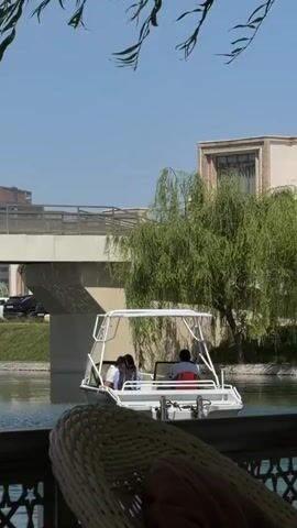 A daytime video shows a small white motorboat with a canopy slowly moving on a calm waterway under a clear, sunny sky. There are several people visible in the boat, one of whom appears to be steering. In the foreground, out of focus, is the arm and part of the legs of a person sitting in a white wicker chair, suggesting they are relaxing by the water.

In the background, a modern-looking bridge spans the water, and beyond it, there are buildings and lush green trees, including a prominent weeping willow. The setting appears to be an urban or park-like area with developed infrastructure, likely a canal or river in Kuchtumgali, Uzbekistan. The overall atmosphere is peaceful and bright. No visible text is present.