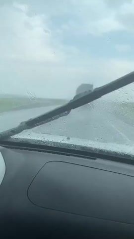 The video shows a view from inside a vehicle looking out through a rain-streaked windshield. In the distance, a large truck is visible on a wet road. The weather appears to be overcast and rainy, with the windshield wipers likely in use, as indicated by the streaked appearance of the glass. The road is lined with green fields on either side, suggesting a rural or semi-rural setting. The dashboard of the vehicle is partially visible in the foreground. The overall impression is one of driving in difficult weather conditions.