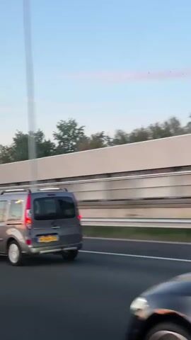 The video shows a black sedan driving on a highway. The sedan, identified by its license plate GGJ-47-V, passes a silver minivan on the right. A sound barrier runs parallel to the road, with trees visible behind it. The sky is light blue with a few clouds. The camera is positioned inside a moving vehicle, tracking the sedan.