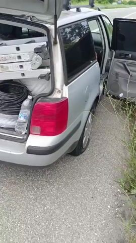 The video begins by showing the open trunk of a silver station wagon parked on the shoulder of a highway. The trunk is filled with various items, including a folded ladder, a cardboard box, a coil of black cable, two orange objects that appear to be filters or air fresheners, a gray bag, and a bottle of water. The camera then pans to the right, revealing a highway with a metal guardrail and trees lining the side. In the distance, another car is seen driving on the highway. The video continues to show the highway from a low angle, with the road and guardrail in focus. Finally, the camera pans up to reveal a wider view of the highway with trees in the background and a cloudy sky above. A person is seen walking on the side of the road, and another car is driving on the highway.