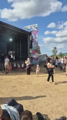 A group of people are gathered outdoors at a festival or concert. In the background, there is a stage with a band performing. People of all ages are present, some are dancing and enjoying the music, while others are standing and watching. The weather appears to be pleasant with a blue sky and some clouds. The ground is dry and dusty, suggesting a summer day.