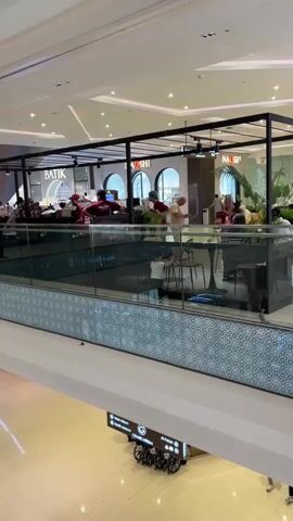 The video shows an overhead view of a modern, multi-level shopping mall interior. In the upper section of the frame, there is a restaurant or cafe area with an open-air design, enclosed by a black metal frame and glass panels. Inside this area, several patrons are seated at tables, some eating and others conversing. The establishment appears to have a contemporary aesthetic with modern furniture and lighting. Signs visible within the establishment include "BATIK" and "NASHI." Below this elevated dining area, the lower level of the mall is visible, featuring a wide open space with a tiled floor. A directional signpost indicating an accessibility icon and possibly store directories is present in the lower mid-ground. A few shoppers are also visible on the lower level, walking. The overall atmosphere suggests a busy but organized commercial environment.