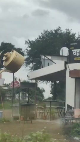 The video shows an outdoor scene with a large, gold-colored teapot sculpture pouring liquid into a white bucket. The teapot is suspended by a red pole. In the background, there are trees and a building. There are also some tables and chairs set up, suggesting an outdoor seating area. The ground is covered with grass and gravel. The sky is overcast.