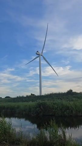 A single wind turbine stands tall against a blue sky with scattered clouds. The turbine has three blades, two of which are clearly visible, and the third is positioned behind the main body of the turbine. The turbine is located on a grassy hill. In the foreground, there is a body of water, likely a pond or a slow-moving river, reflecting the sky and the surrounding vegetation. The vegetation consists of tall green grass and reeds. The overall scene depicts a serene natural landscape with a prominent man-made structure for renewable energy generation.
