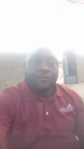 The video shows a person with dark skin wearing a red polo shirt with "The Hope Institute of Canada" embroidered on it. They are speaking in the video. The background is blurry, but it appears to be an outdoor setting with trees and possibly a brick structure. The video's lighting fluctuates between bright and dark, possibly due to changing natural light conditions.
