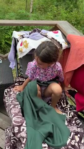 A young girl with dark hair is crouching on a deck, surrounded by laundry. She is wearing a pink t-shirt with a patterned design and a dark green skirt. She is partially covered by a large, brown and white patterned blanket and a green blanket. In the background, there is a wooden railing, trees, and a table with various items on it, including a doll and sunglasses. Another person's voice calls out to the girl. The scene appears to be outdoors, possibly in a backyard or a cabin setting.