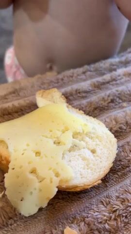A close-up shot shows a hand reaching for a slice of bread topped with melted cheese. The bread is a light golden brown, and the cheese is a pale yellow with some translucent areas and small holes, suggesting it is a semi-hard cheese. The cheese is slightly melted and appears soft and gooey. The bread and cheese are placed on a brown, textured surface, possibly a towel or blanket. In the blurred background, a small child's torso is visible, suggesting the scene is indoors and likely a casual mealtime. The lighting appears to be natural and soft, indicating it could be daytime. The focus is on the food and the action of taking a bite. There is no visible text in the frame. The overall impression is one of simple, homemade comfort food being prepared or enjoyed.