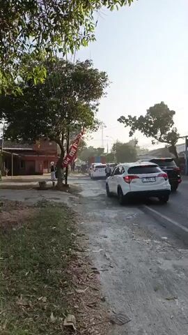 The video shows a road with several vehicles, including cars and motorcycles. A white SUV is in the foreground, with a motorcycle passing it on the right. Several other cars and motorcycles are visible in the background, moving in the same direction. The road appears to be made of asphalt and is partially covered in gravel and dirt. There are trees and buildings on the left side of the road, and a power pole with wires is visible in the distance. The sky is clear and bright, indicating daytime. A person is standing on the left side of the road, near a red flag. Two people on a motorcycle are visible in the middle ground, with the passenger wearing a headscarf. The overall impression is of a busy street scene with moderate traffic.