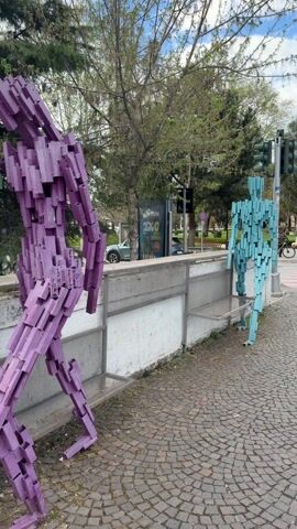 The video shows three colorful human-like sculptures made out of small rectangular blocks. The sculptures are painted purple, green, and turquoise. One is standing, another is seated on a low wall, and the third is out of frame on the right side. Behind the sculptures is a canal of flowing water, framed by green trees and grass. There are pink blossoming trees in the distance. The sky is partly cloudy with patches of blue. The camera pans around the sculptures and environment.