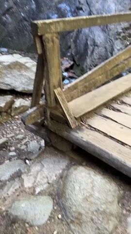 The video shows a weathered wooden bridge with a railing overlooking a narrow stream at the base of a rocky cliff. The bridge is made of rough-hewn planks and appears to be in disrepair. The stream flows over rocks and into a pool at the bottom of a waterfall. The waterfall cascades down a tall, sheer rock face, surrounded by moss and vegetation. The lighting is dim, suggesting an overcast day or a shaded area. The camera moves slowly, panning across the scene from the perspective of someone standing on the bridge.
