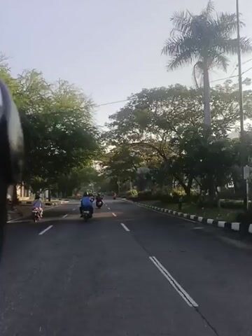 A first-person perspective video shows a road with several motorcycles riding ahead. The road is lined with trees and has white dashed lines marking the lanes. The sun is visible in the sky, creating a bright atmosphere. A green street sign is visible at the 00:03 mark, indicating directions to various locations. The video appears to be taken during the daytime.