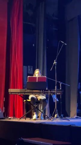 A young woman sits at an electronic keyboard on a stage, facing the audience. She is wearing a light-colored hoodie and dark pants. Her hands are on the keyboard, and she appears to be playing or singing. A music stand with a red binder is positioned in front of her. Two microphones are set up on stands near her. Red curtains hang to her left, and blue curtains are visible behind her. The stage floor is dark.