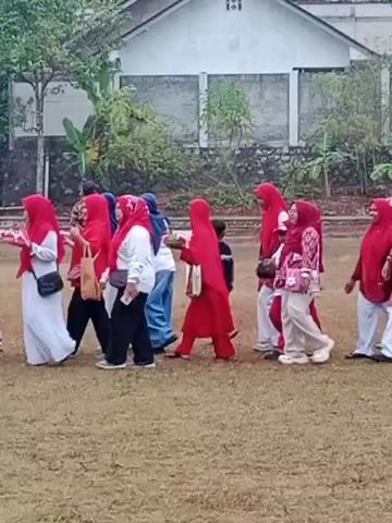 A group of people, mostly women in hijabs and men and children, are walking across a grassy field. Some of the women are wearing red hijabs and matching outfits. One woman is holding a baby. The people appear to be walking in a line or procession. In the background, there is a building and some trees. The ground is covered with dry grass. The overall impression is of an outdoor gathering or event.