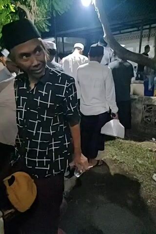 A group of men are gathered outdoors at night. The scene is illuminated by a single light source hanging from a tree. Many of the men are wearing traditional Islamic clothing, including kufis and sarongs. One man in the foreground is wearing a black and white checkered shirt and a black kufi. He is looking towards the camera and holding a small, metallic object. Other men are seen walking, some carrying plastic bags. The ground appears to be dirt and there are some plants and trees visible. The overall impression is of a community gathering or event taking place after dark.