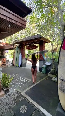 A woman in a black swimsuit and barefoot carries a surfboard on her head, walking away from the camera toward a wooden gate. The gate is adorned with the sign "MEDEWI BEACH INN." To the left of the gate are several surfboards leaning against a wall. The area is landscaped with trees, potted plants, and decorative stones. A motorcycle is parked near the entrance, and a green trash can stands to the right of the gate.