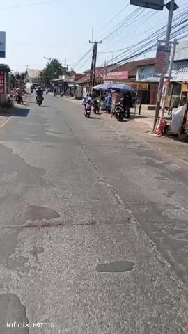 The video shows a street scene from the perspective of someone on a motorcycle. In the foreground, the handlebars and a portion of the motorcycle's mirror are visible. The street is paved and appears somewhat worn. On the right side of the street, there are several small businesses or stalls. One stall has an umbrella and appears to be selling food, with people gathered around it. Another stall is a cart with wheels, also under an umbrella. Further down the street, there are more buildings and stalls, including one with a sign that says "Tahu Goreng" (fried tofu). On the left side of the street, there are houses and more shops. Several motorcycles are parked or being ridden on the street. There are also power lines and utility poles overhead. The sky is clear and the sun is shining, indicating it is daytime. The overall impression is a busy street in a commercial or residential area.