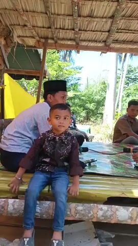 A young boy in a patterned shirt and jeans sits facing forward, looking slightly to his right. He is positioned on a raised platform covered with a yellowish material. Behind him, a man in a blue and white striped shirt and a black cap is visible. To the right of the boy, another man in a brown shirt sits facing away from the camera. Further to the right, another person in a black shirt and patterned sarong is seated and looking down. Microphones are present on the platform. The scene appears to be outdoors, under a thatched roof structure, with lush green foliage and trees in the background. The lighting suggests it is daytime.
