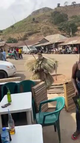 The video shows a person dressed in a full-body costume made of dry grass or straw standing on a road in a village or small town. The costume covers the person completely, making them appear as a large, amorphous, textured figure. The person holds a thin stick or branch. The background includes a road with a van and motorcycle passing by, buildings with corrugated metal roofs, and a hill in the distance. In the foreground, there are plastic chairs and tables, suggesting an outdoor eating area. Bottles of drinks are visible on the table. The camera is handheld and slightly shaky. A person in the foreground is handling a green bag. There is laughter in the background.
