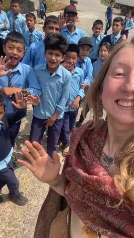 A blonde woman takes a selfie video with a large group of kids in school uniforms. The children are all smiling and waving. The woman is wearing a colorful scarf. The background shows a road and some buildings. There is a white truck parked in the distance. The lighting is bright and sunny.