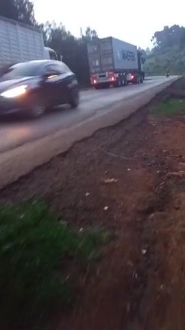 The video shows a rural roadside scene, featuring several trucks and a motorcycle on a paved road. The camera is positioned off the road, on a muddy, uneven shoulder. There are trees visible in the background and on the hillside. The road has cracks and a somewhat damaged edge, indicating wear. A few trucks with illuminated taillights pass by, heading up a slight incline. A person on a motorcycle is seen traveling the road in the same direction as the trucks. The overall lighting suggests it is either early morning or late afternoon with overcast weather.