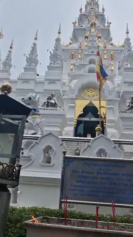 The video shows a white temple with golden accents and a flag in front of it. There is a sign in front of the temple with Thai writing on it. The sign has incense sticks in front of it. There is a flame on the incense sticks, indicating they are lit. The temple has multiple tiers and ornate decorations.