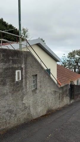 The camera pans up an asphalt road running downhill past houses on both sides. A concrete wall with a railing on top runs along the left side of the road. The wall supports one side of a light yellow house with a dark red roof. Across the road to the right is a stone wall and a locked iron gate, giving way to trees and vegetation. In the distance, the road goes down into a city and the ocean is visible. Above, the sky is overcast.