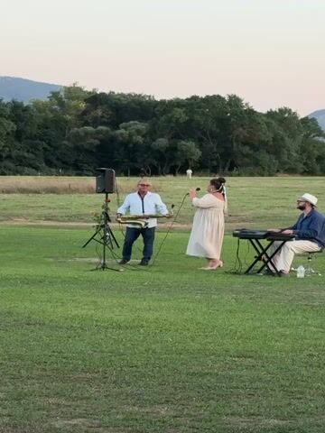 A trio performs music outdoors. A man in a white shirt and blue jeans stands center left, holding a saxophone and wearing sunglasses. To his right, a woman in a long, light-colored dress sings into a microphone. To the right of the woman, a man in a blue shirt and light-colored pants sits at a keyboard, wearing a hat. They are on a grassy field with trees and hills in the background. A speaker on a stand is positioned next to the saxophonist.