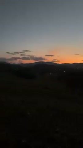 The video shows a landscape at dusk, likely in a rural or mountainous area. The sky is a gradient of colors from dark blue above to orange and pink near the horizon, suggesting either sunrise or sunset. There are some clouds scattered across the sky. In the distance, there are silhouettes of mountains, and some lights are visible indicating a small town or settlement. The foreground is mostly dark and appears to be a field or grassy area. The overall mood is serene and peaceful.