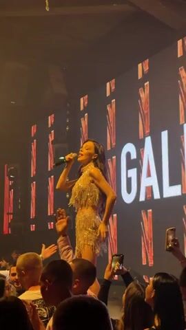 A female singer with long wavy hair, wearing a sparkling gold fringed outfit, performs on a raised platform in front of a large digital screen displaying the word "GALINA". She holds a microphone in her right hand. The audience surrounds the stage, with many people holding up their phones to record her performance. Bright spotlights shine on the singer from above. The environment appears to be a nightclub or concert venue.