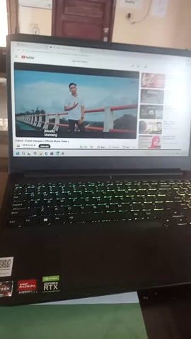 The video opens with a close-up of a laptop screen, displaying a YouTube video. The video within the YouTube video shows a young man in a white shirt standing outside, looking to his left. The background appears to be a bridge with water visible below. The YouTube interface shows the video title "Zahira - Putih (Official Music Video)". The laptop keyboard is visible on the right side of the screen, with its backlight illuminating the keys in a purplish hue. To the left of the laptop, a wooden door and a whiteboard are visible. The lighting is dim, with a warm glow emanating from the laptop screen.