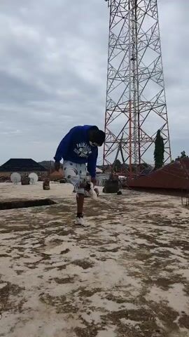 A person is standing on a rooftop with a large communication tower in the background. The person is wearing a blue hoodie, white shorts with a pattern, and white sneakers. They also have goggles on. The sky is cloudy. The rooftop appears to be made of concrete and has some debris on it. There are some buildings in the distance. The person is looking down at the ground.