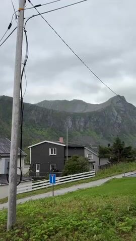 A utility pole with wires stands in the foreground of a video that pans across a residential area with mountains in the background. The sky is overcast. The first house visible is dark gray with a white fence. Next is a white house with a gray roof and a wooden garage door. A person is riding a bicycle on a road in front of the houses. A body of water is visible behind some houses and to the left of the mountains.