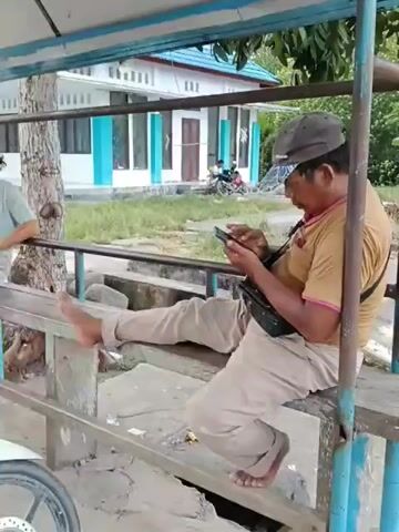 The video shows two individuals sitting on a wooden bench under a sheltered structure. The person on the right is a man wearing a brown t-shirt, tan pants, and a dark baseball cap. He is barefoot and is actively using a mobile phone, holding it with both hands and looking down at the screen. He has a black bag slung across his chest. The person on the left is partially visible, wearing a light blue t-shirt and dark pants. This individual is also looking down at a mobile phone, which they are holding with one hand. Their legs are also extended. In the background, there is a white building with blue accents, and some trees. A motorcycle is parked in the foreground on the left side of the frame. The ground appears to be dirt or sand, with some debris scattered around. The overall setting seems to be outdoors, possibly a rest area or a waiting area.