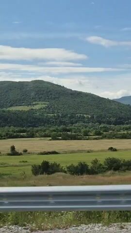 The video shows a view from a moving vehicle. The scene is a rural landscape with rolling green hills and fields. There is a metal guardrail in the foreground. The sky is blue with scattered white clouds. The terrain is uneven, with some trees and vegetation visible on the slopes of the hills.