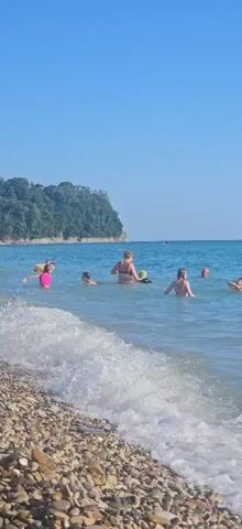 A group of people are swimming in the ocean on a sunny day. The water is blue and clear, with waves breaking on a pebble beach in the foreground. A woman in a red bikini is standing in the water, holding a green object. Several children are swimming around her. In the background, a green cliff covered in trees slopes down to the sea. The sky is clear and blue.